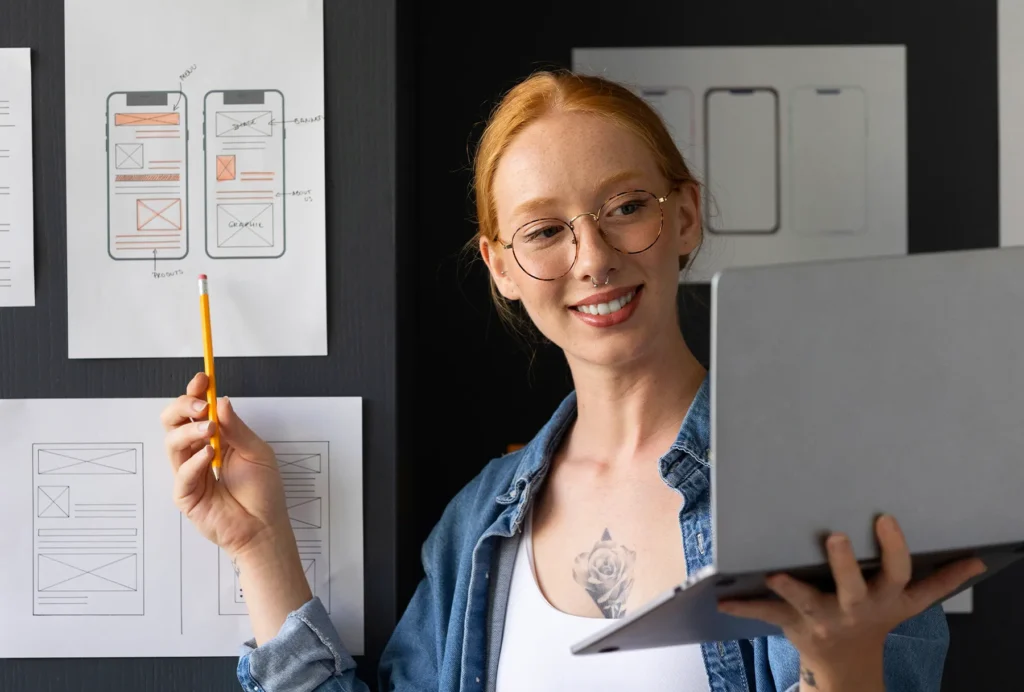 A person holding up a laptop while pointing to wireframes on a whiteboard