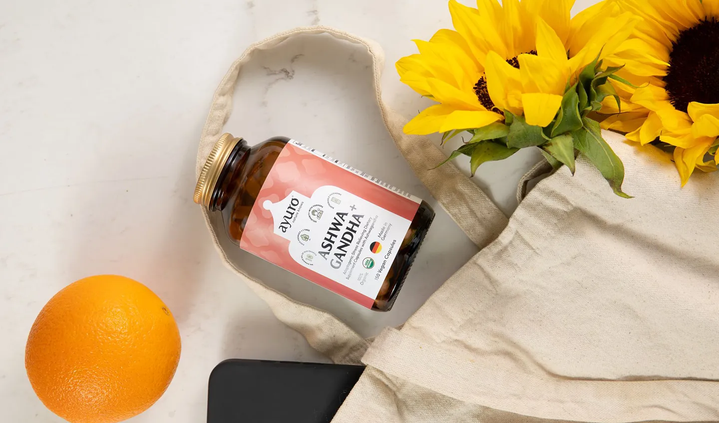 A bottle of ayuro Ashwagandha lays on a marble countertop with an orange, a tote bag and sunflowers next to it